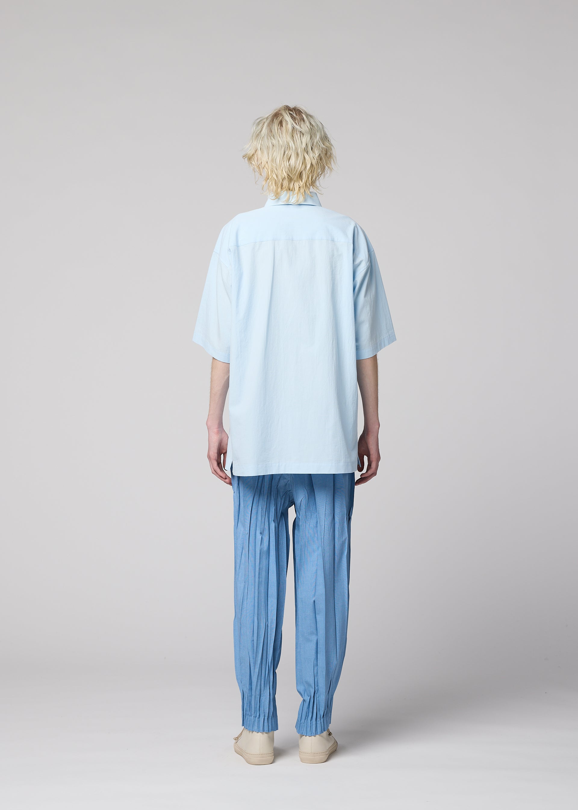 Zoomed view A person with curly blond hair stands facing away, wearing the IM MEN WHITE SHIRT—a loose, short-sleeved cotton linen ramie top—paired with pleated blue pants gathered at the ankles and beige shoes, set against a plain light gray background.