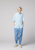 A person with wavy blond hair wears the WHITE SHIRT from IM MEN, a loose, light blue cotton linen ramie shirt, paired with pleated blue pants featuring gathered ankles and beige shoes, standing against a plain light gray background.
