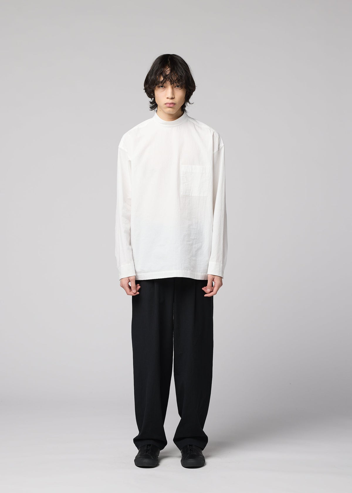 A person with medium-length dark hair stands facing forward against a plain light gray background, wearing the IM MEN WHITE SHIRT— a loose white cotton-linen, long-sleeve top — paired with black wide-leg pants and black shoes. The relaxed silhouette of the outfit adds to its effortless appeal. view 6 of 8