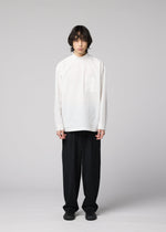 A person with medium-length dark hair stands facing forward against a plain light gray background, wearing the IM MEN WHITE SHIRT— a loose white cotton-linen, long-sleeve top — paired with black wide-leg pants and black shoes. The relaxed silhouette of the outfit adds to its effortless appeal.