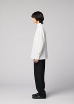 A person with medium-length dark hair stands in profile against a plain light gray background, wearing the IM MEN WHITE SHIRT—a loose, long-sleeved pullover with a relaxed silhouette—paired with black wide-leg pants and black shoes.