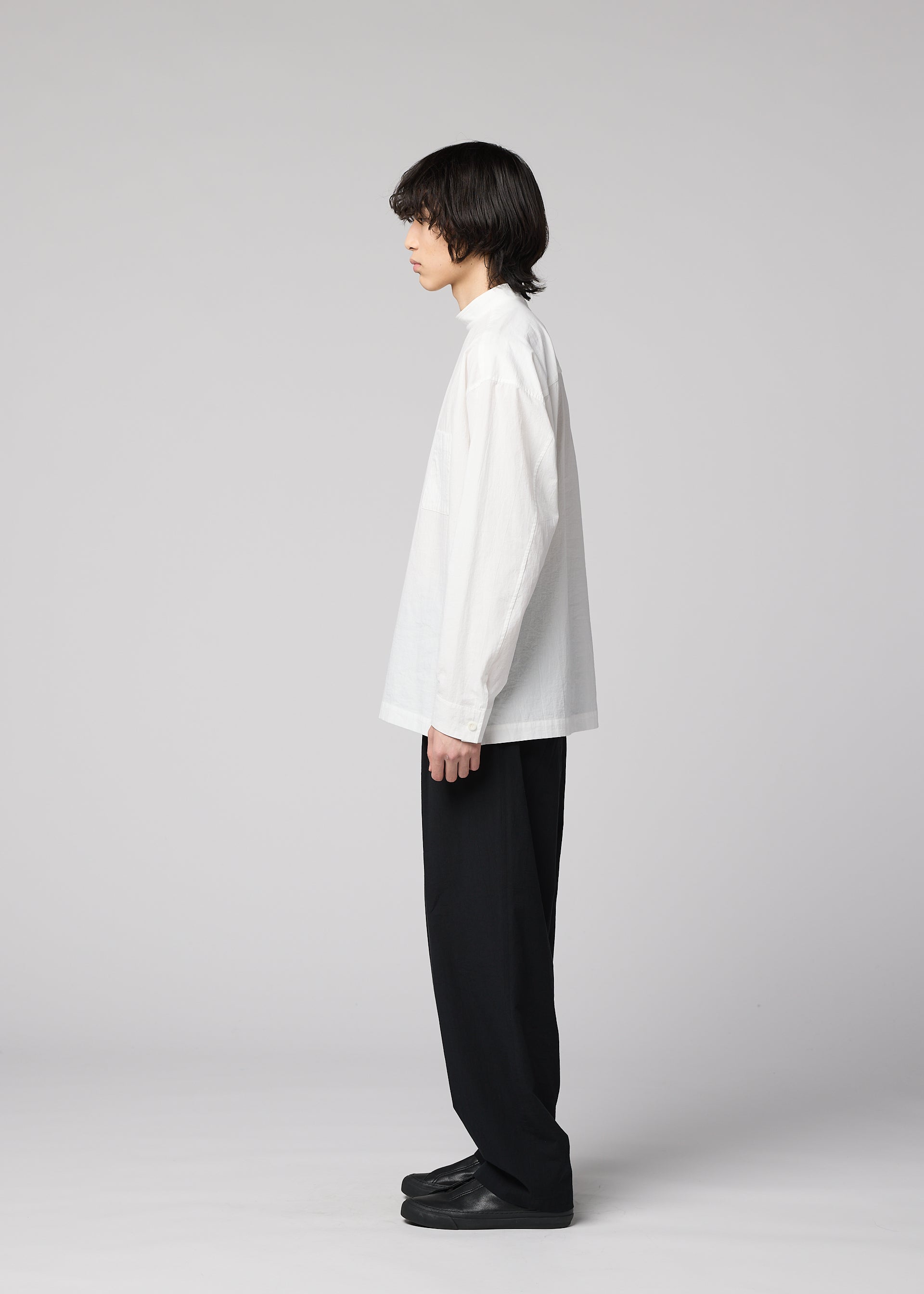Zoomed view A person with medium-length dark hair stands in profile against a plain light gray background, wearing the IM MEN WHITE SHIRT—a loose, long-sleeved pullover with a relaxed silhouette—paired with black wide-leg pants and black shoes.
