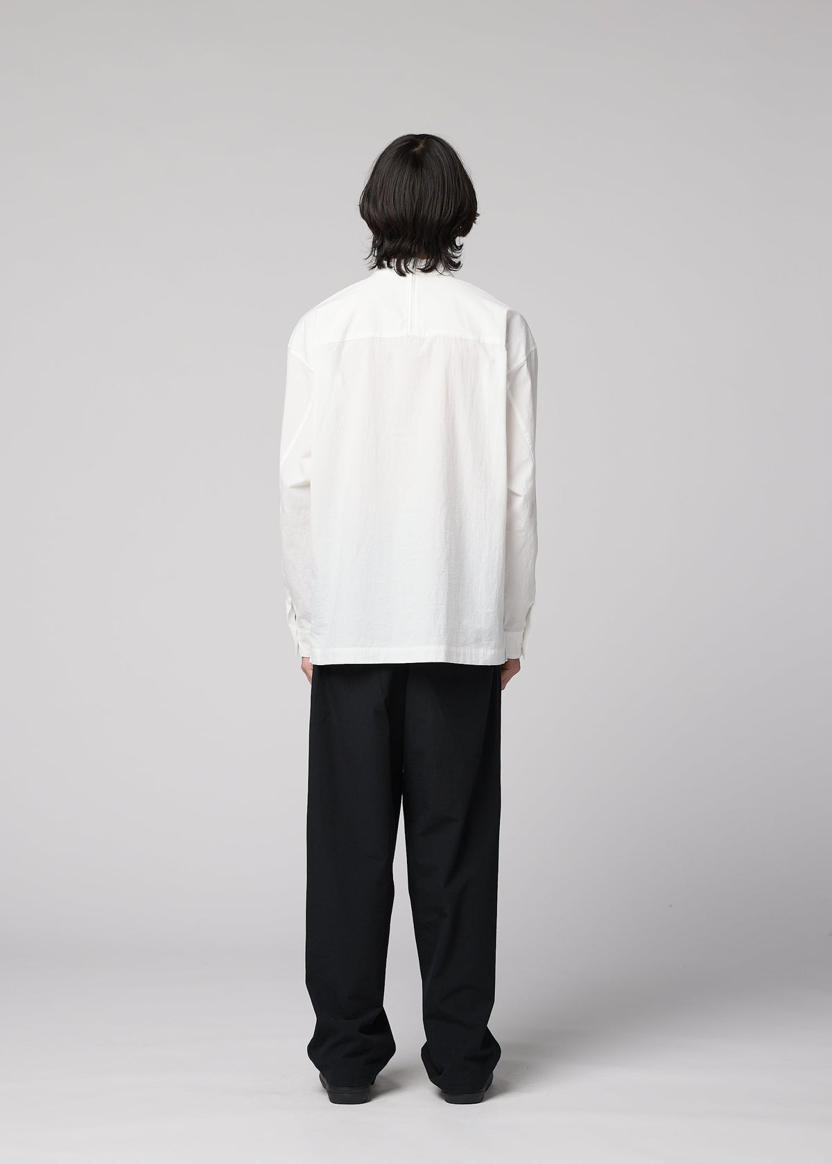 An individual with medium-length dark hair stands facing away, wearing the IM MEN WHITE SHIRT— a white, long-sleeved pullover made from cotton linen—paired with loose-fitting black pants featuring a relaxed silhouette, set against a plain light gray background. view 8 of 8