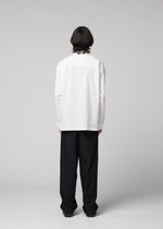 An individual with medium-length dark hair stands facing away, wearing the IM MEN WHITE SHIRT— a white, long-sleeved pullover made from cotton linen—paired with loose-fitting black pants featuring a relaxed silhouette, set against a plain light gray background.