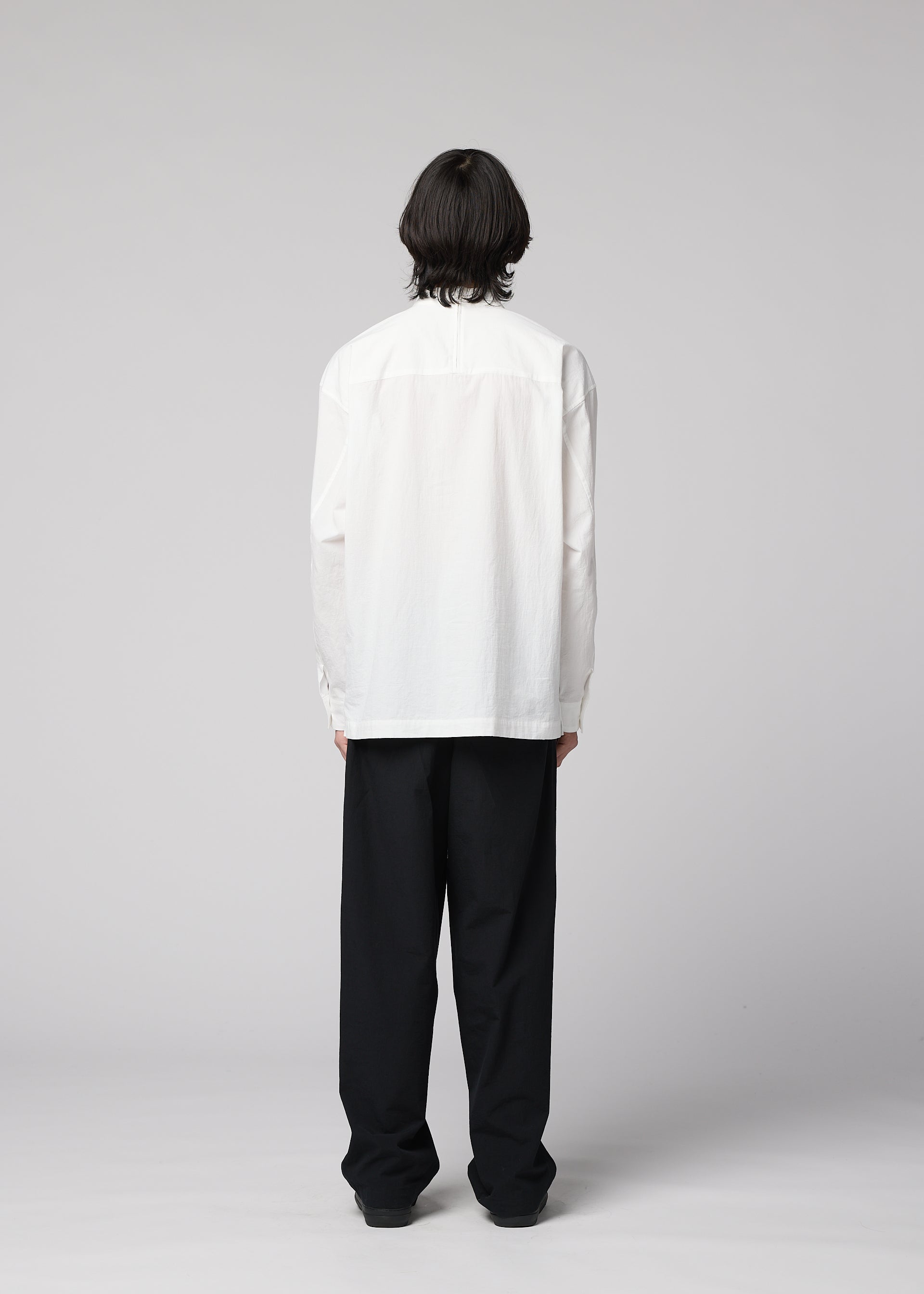Zoomed view An individual with medium-length dark hair stands facing away, wearing the IM MEN WHITE SHIRT— a white, long-sleeved pullover made from cotton linen—paired with loose-fitting black pants featuring a relaxed silhouette, set against a plain light gray background.
