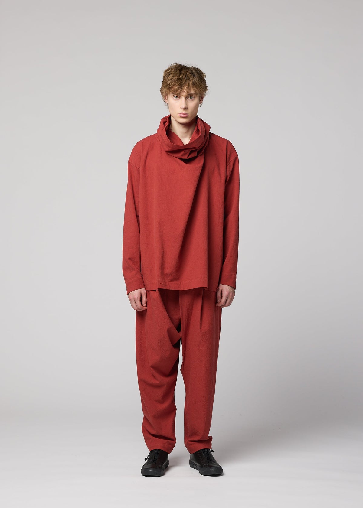 A person with short, light brown hair stands facing forward, dressed in the IM MEN COTTON YAK set—a loose-fitting red ensemble crafted from premium cotton yak fabric with a draped neckline and matching pants. Inspired by the IM MEN Autumn Winter 2026/27 collection, their look is completed with black shoes and photographed against a plain light gray background. view 7 of 11