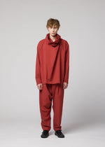 A person with short, light brown hair stands facing forward, dressed in the IM MEN COTTON YAK set—a loose-fitting red ensemble crafted from premium cotton yak fabric with a draped neckline and matching pants. Inspired by the IM MEN Autumn Winter 2026/27 collection, their look is completed with black shoes and photographed against a plain light gray background.