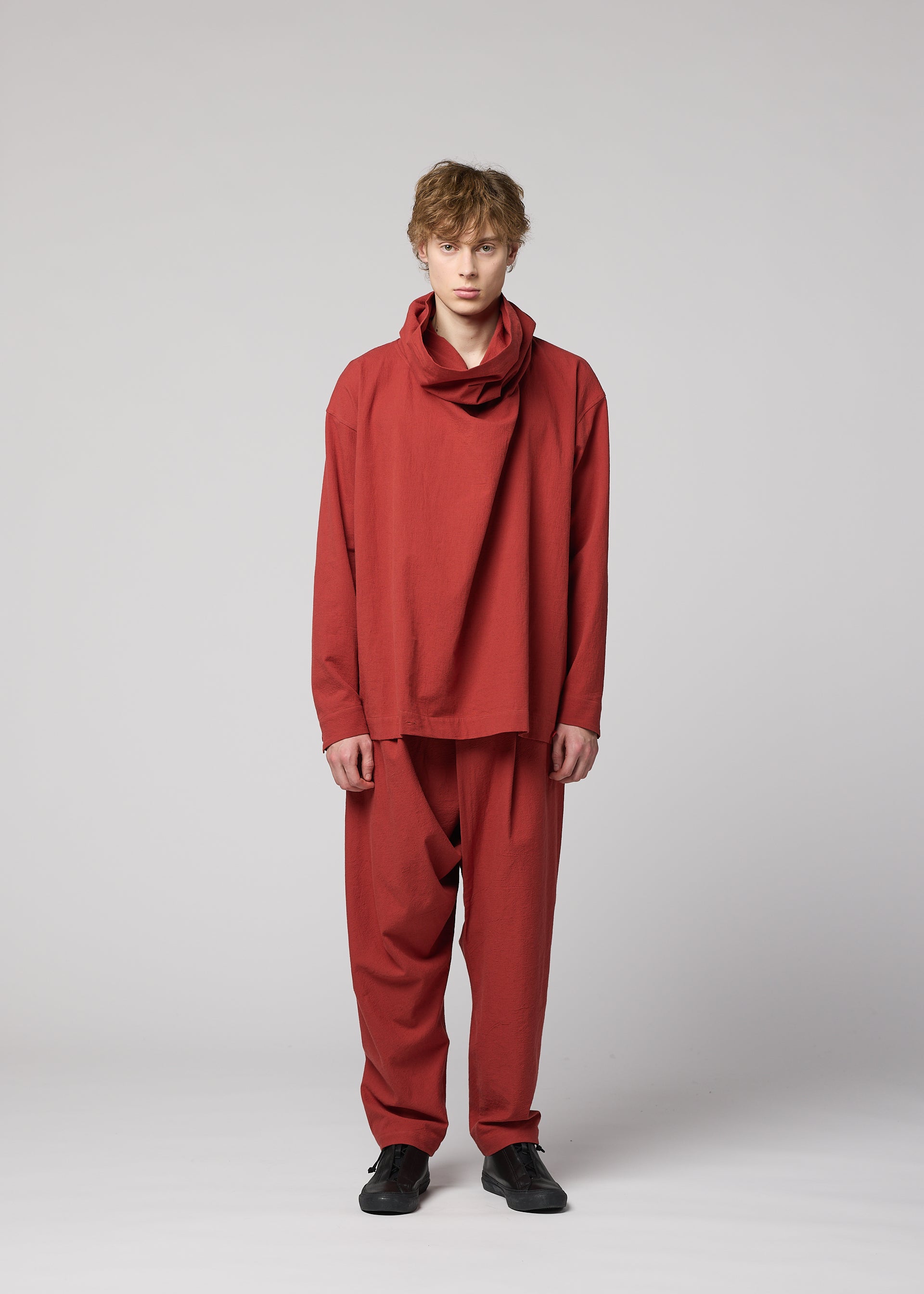 Zoomed view A person with short, light brown hair stands facing forward, dressed in the IM MEN COTTON YAK set—a loose-fitting red ensemble crafted from premium cotton yak fabric with a draped neckline and matching pants. Inspired by the IM MEN Autumn Winter 2026/27 collection, their look is completed with black shoes and photographed against a plain light gray background.