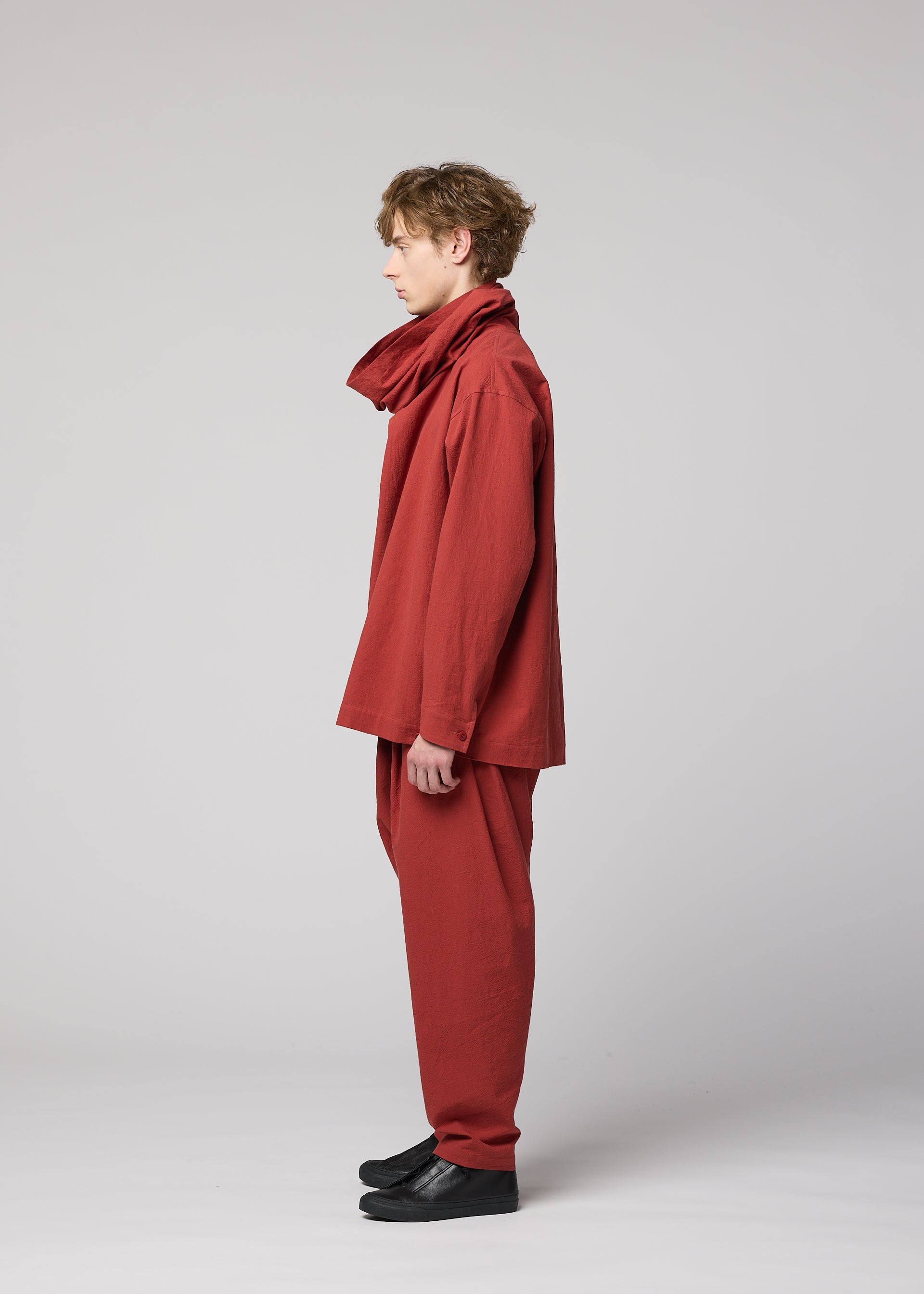 Zoomed view A person stands in profile against a plain background, wearing loose-fitting, matching red pants and a long-sleeved high-neck shirt crafted from IM MEN’s COTTON YAK fabric. Their outfit is completed with a wide scarf and black shoes, their curly hair adding to the understated elegance inspired by IM MEN Autumn Winter 2026/27.