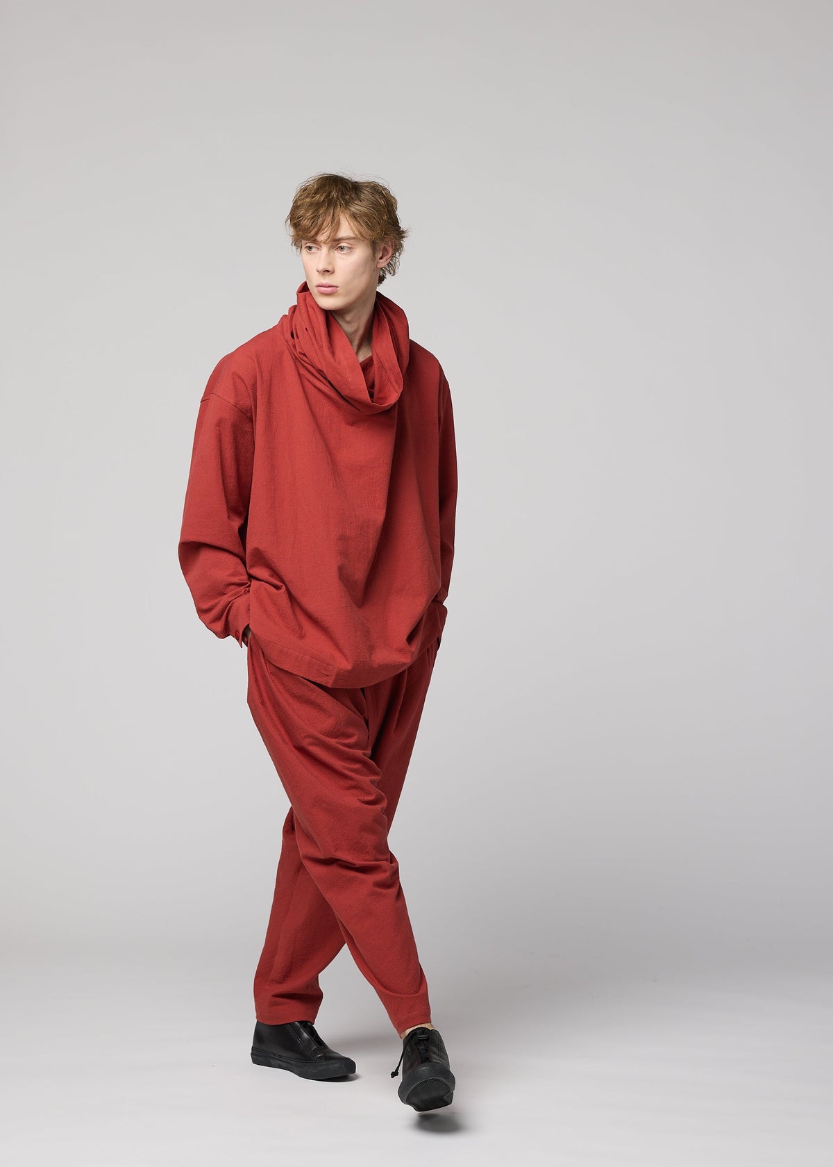A person stands against a plain light gray background, hands in pockets and looking to the side, dressed in a loose-fitting COTTON YAK outfit from IM MEN, featuring soft cotton yak fabric in rust color with a draped collar—a modern look inspired by the Autumn Winter 2026/27 collection. view 10 of 11