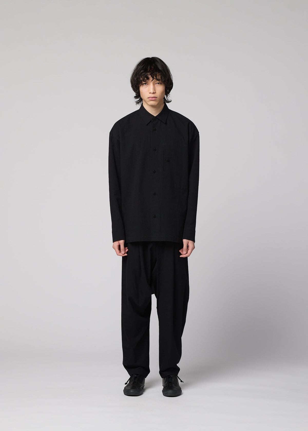 A person with medium-length dark hair stands against a plain gray background, wearing a loose-fitting IM MEN COTTON YAK shirt featuring a natural texture, paired with wide black pants and black shoes, facing forward with a neutral expression. view 6 of 9