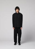 A person with medium-length dark hair stands against a plain gray background, wearing a loose-fitting IM MEN COTTON YAK shirt featuring a natural texture, paired with wide black pants and black shoes, facing forward with a neutral expression.