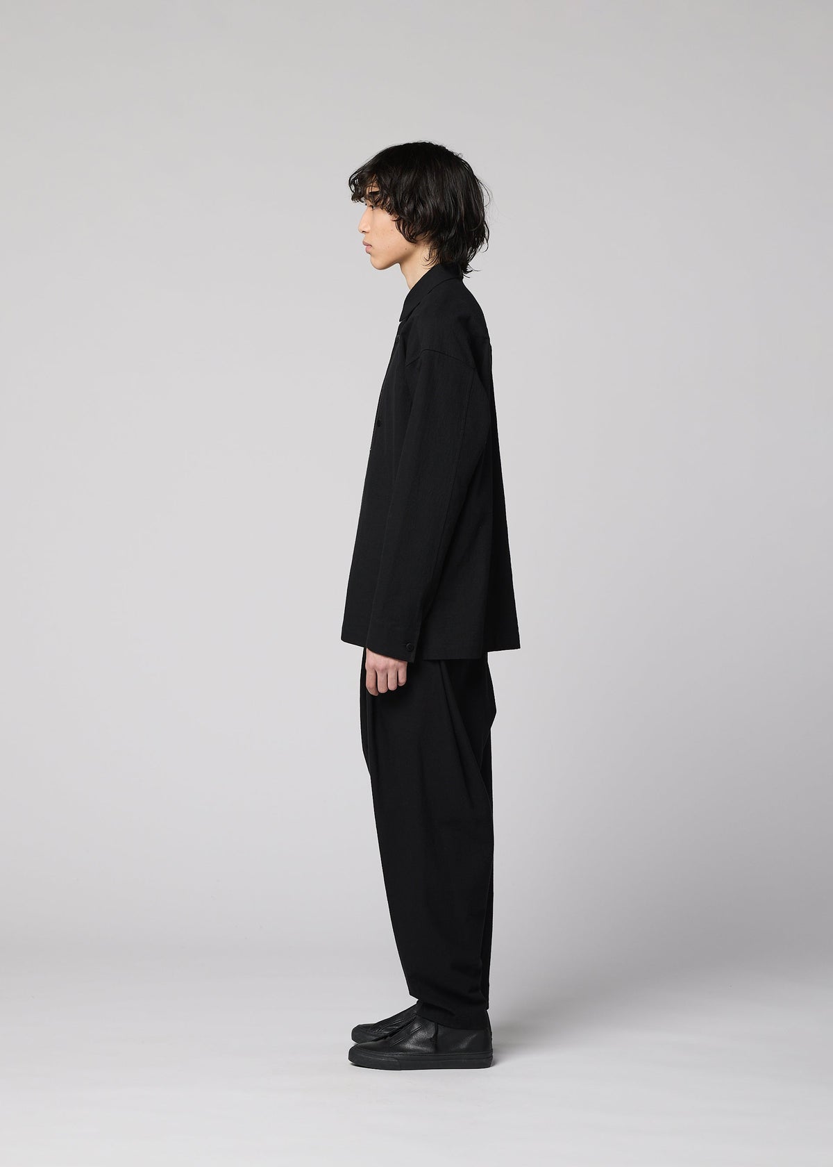 A person with medium-length dark hair stands in profile against a plain light gray background, wearing loose-fitting black pants, black shoes, and the IM MEN COTTON YAK long-sleeved shirt that highlights its natural texture. view 7 of 9