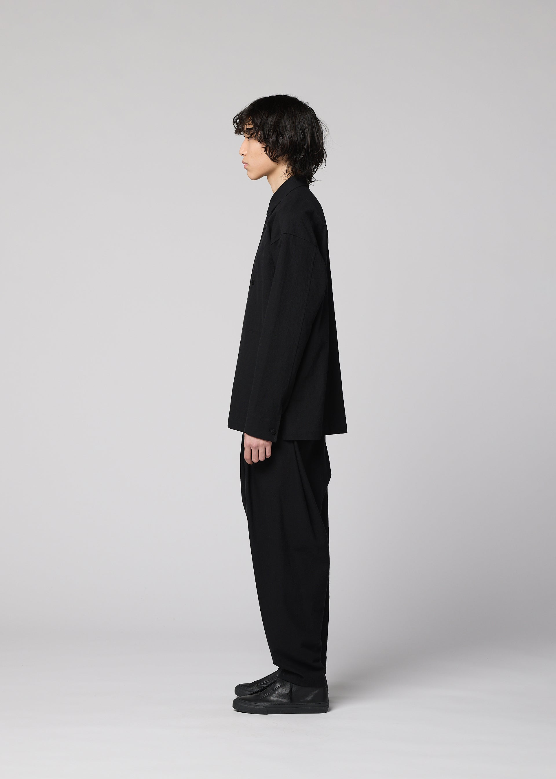 Zoomed view A person with medium-length dark hair stands in profile against a plain light gray background, wearing loose-fitting black pants, black shoes, and the IM MEN COTTON YAK long-sleeved shirt that highlights its natural texture.