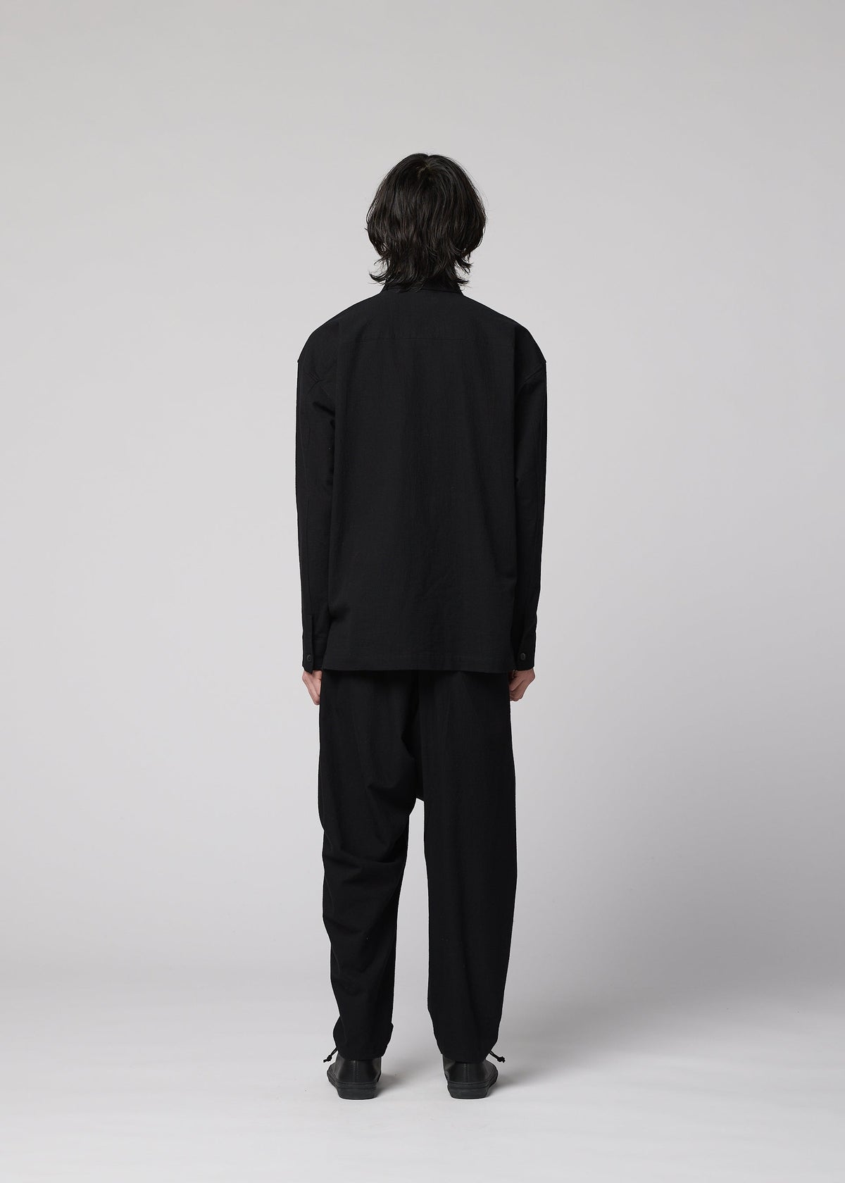 A person with shoulder-length dark hair stands facing away, wearing the IM MEN COTTON YAK loose black long-sleeved shirt made from natural-texture cotton yak fabric, paired with black trousers and black shoes, against a plain light gray background. view 8 of 9