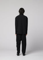 A person with shoulder-length dark hair stands facing away, wearing the IM MEN COTTON YAK loose black long-sleeved shirt made from natural-texture cotton yak fabric, paired with black trousers and black shoes, against a plain light gray background.