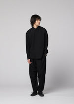 A person with medium-length dark hair stands against a plain light background, wearing the loose-fitting, long-sleeved COTTON YAK shirt from IM MEN, which showcases a natural texture. The shirt is paired with wide black pants and black shoes, as they look slightly to the side.