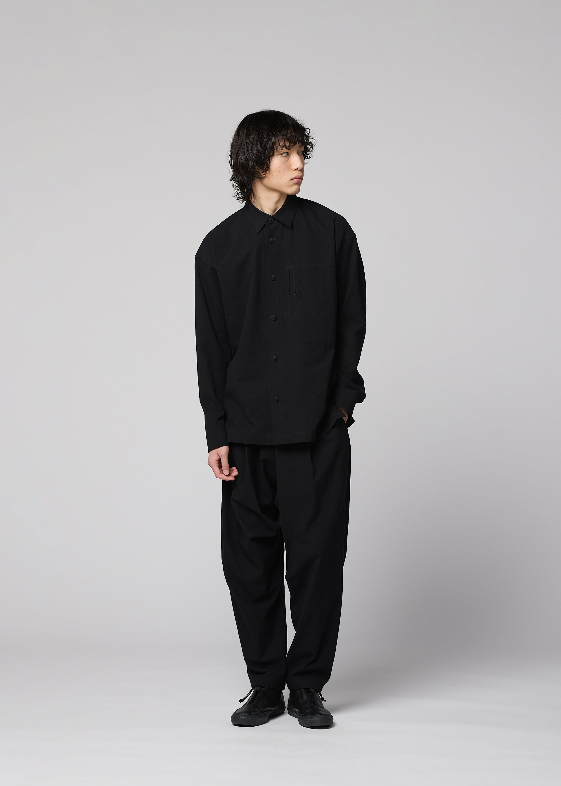 Zoomed view A person with medium-length dark hair stands against a plain light background, wearing the loose-fitting, long-sleeved COTTON YAK shirt from IM MEN, which showcases a natural texture. The shirt is paired with wide black pants and black shoes, as they look slightly to the side.