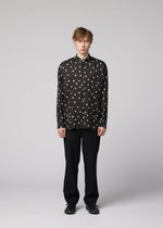 A person with light brown hair stands facing forward, dressed in a black MERRY SATIN SHIRT by IM MEN with white polka dots, paired with black pants and black shoes, against a plain light grey background.