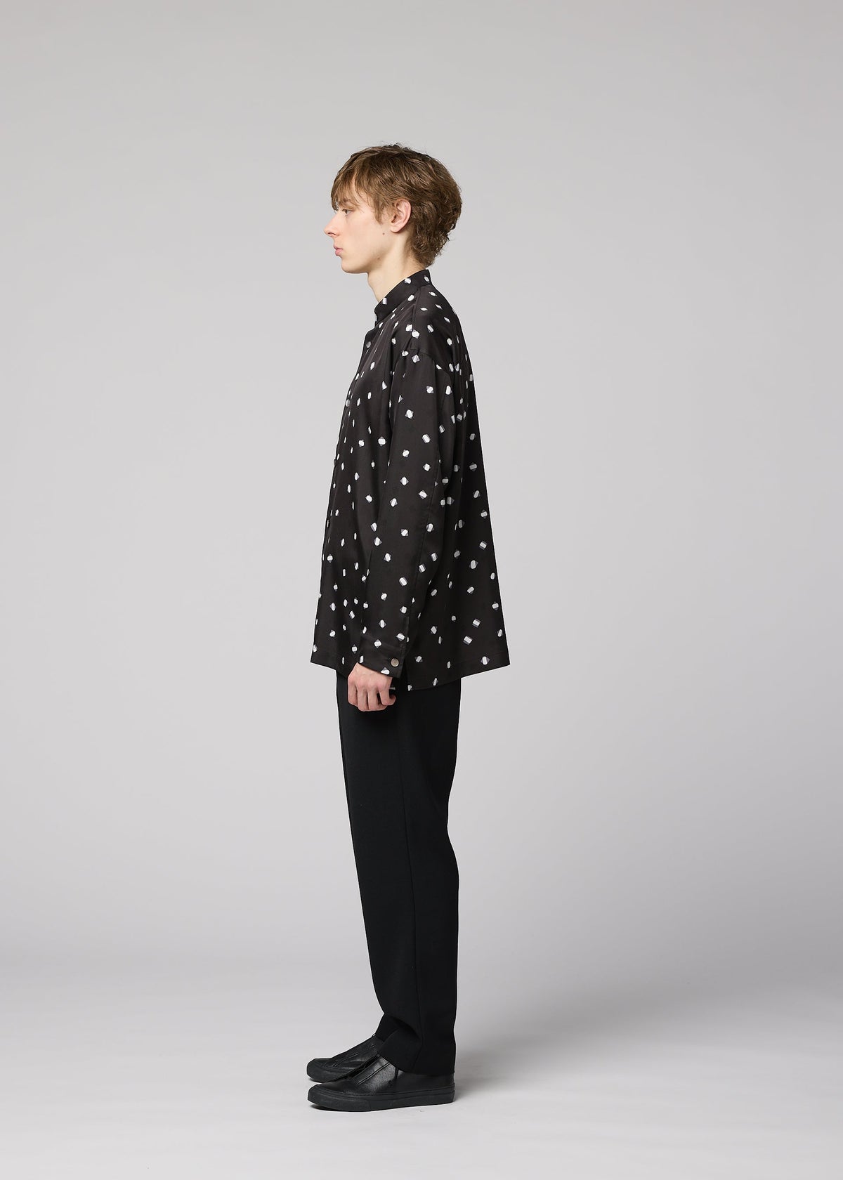 A person stands in profile against a plain background, wearing the IM MEN MERRY SATIN SHIRT—a black shirt with white polka dots from the Autumn Winter 2026/27 collection—paired with black pants and black shoes. view 7 of 9
