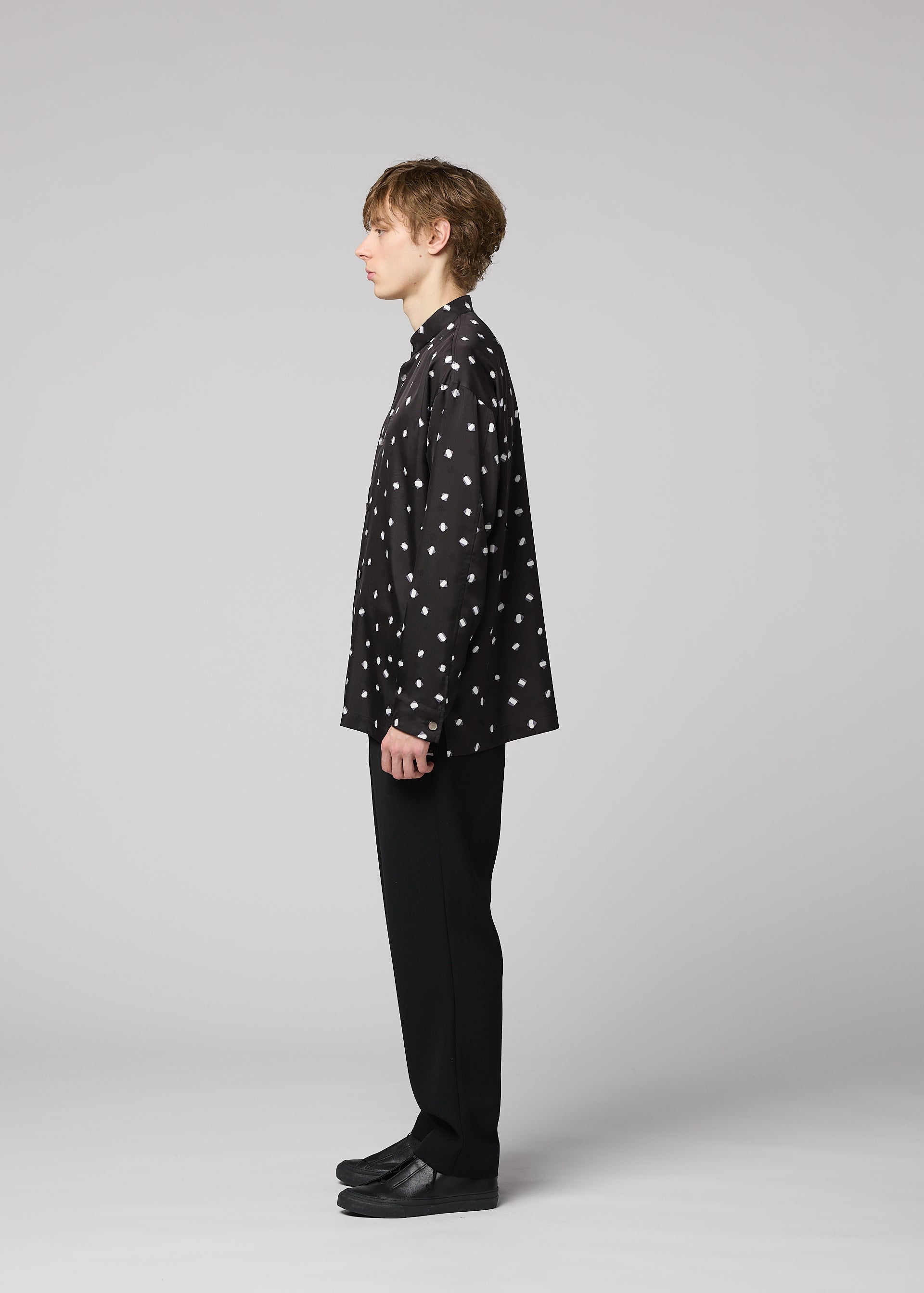 Zoomed view A person stands in profile against a plain background, wearing the IM MEN MERRY SATIN SHIRT—a black shirt with white polka dots from the Autumn Winter 2026/27 collection—paired with black pants and black shoes.