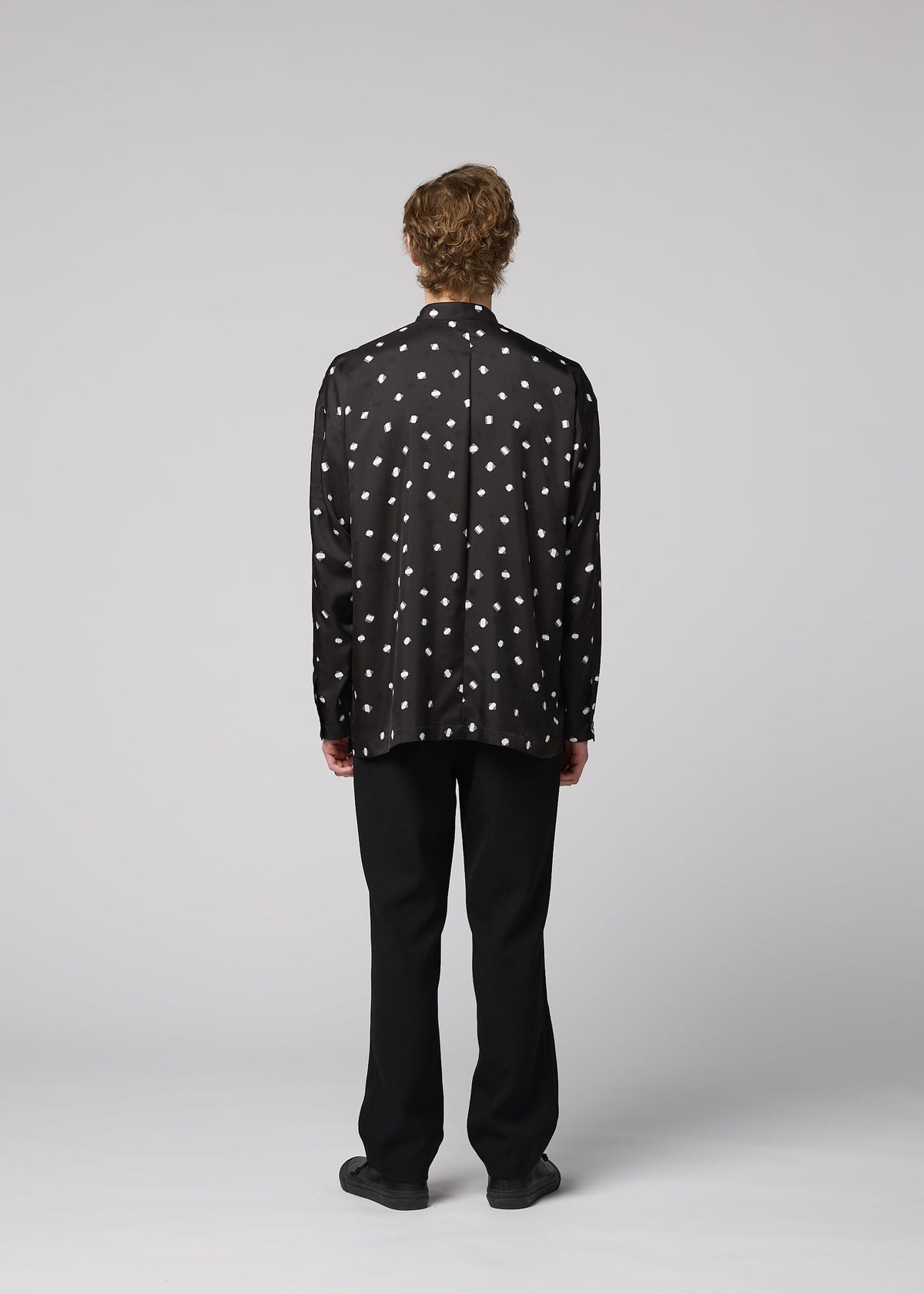 A person with short, curly hair stands facing away, dressed in the IM MEN MERRY SATIN SHIRT—a black satin shirt with white polka dots featuring a stand-up collar—paired with black pants and black shoes, set against a plain light gray background. The ensemble reflects the sleek elegance of IM MEN’s Autumn Winter 2026/27 collection. view 8 of 9