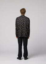 A person with short, curly hair stands facing away, dressed in the IM MEN MERRY SATIN SHIRT—a black satin shirt with white polka dots featuring a stand-up collar—paired with black pants and black shoes, set against a plain light gray background. The ensemble reflects the sleek elegance of IM MEN’s Autumn Winter 2026/27 collection.