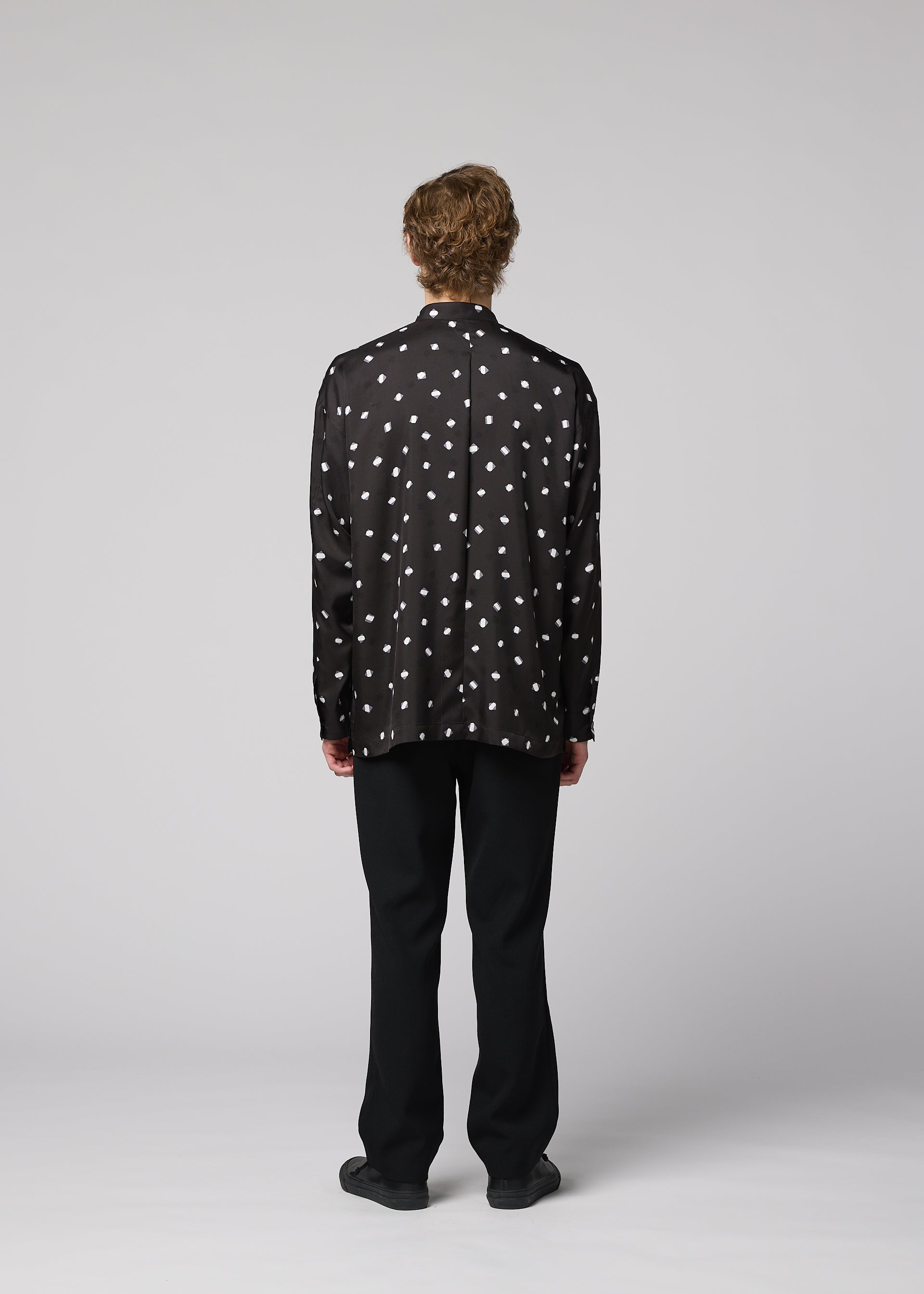 Zoomed view A person with short, curly hair stands facing away, dressed in the IM MEN MERRY SATIN SHIRT—a black satin shirt with white polka dots featuring a stand-up collar—paired with black pants and black shoes, set against a plain light gray background. The ensemble reflects the sleek elegance of IM MEN’s Autumn Winter 2026/27 collection.