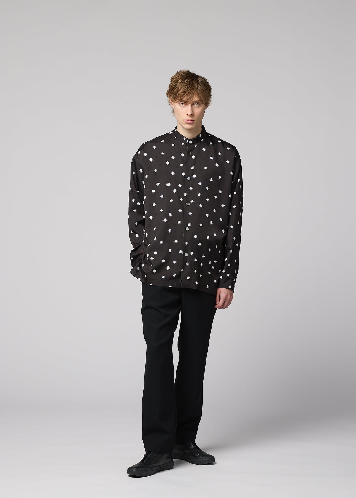 A person stands against a plain light gray background, wearing the IM MEN MERRY SATIN SHIRT—a black button-up shirt with white polka dots from the Autumn Winter 2026/27 collection—paired with black pants and black shoes. Their hands are in their pockets, and they have short, tousled hair. view 9 of 9