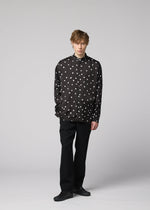 A person stands against a plain light gray background, wearing the IM MEN MERRY SATIN SHIRT—a black button-up shirt with white polka dots from the Autumn Winter 2026/27 collection—paired with black pants and black shoes. Their hands are in their pockets, and they have short, tousled hair.