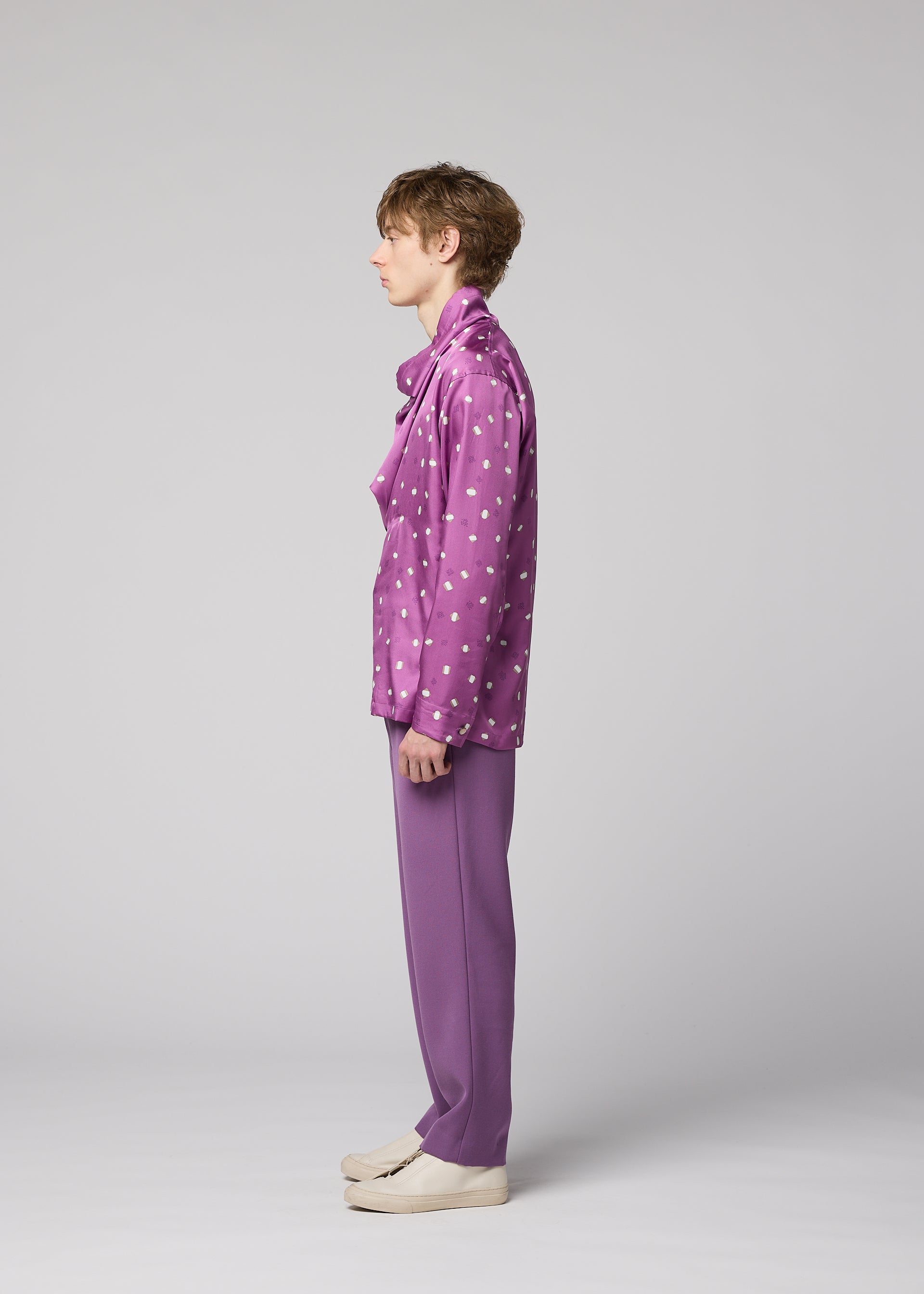 Zoomed view A person stands in profile against a gray background, wearing the MERRY SATIN SHIRT by IM MEN—a shiny satin shirt with white polka dots—paired with matching purple pants and off-white shoes.