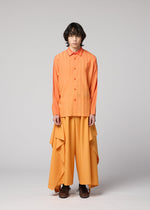 A person with wavy hair wears the DAWN RAY SHIRT by IM MEN, a transfer-printed orange pleated shirt, paired with wide, draped, flowing orange pants that feature a subtle gradation pattern. They stand against a plain gray background with a neutral expression.