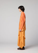 Wearing the IM MEN DAWN RAY SHIRT in orange, a person with medium-length dark hair stands in profile against a plain background, pairing it with a long, flowing yellow skirt featuring a subtle gradation pattern and brown shoes.