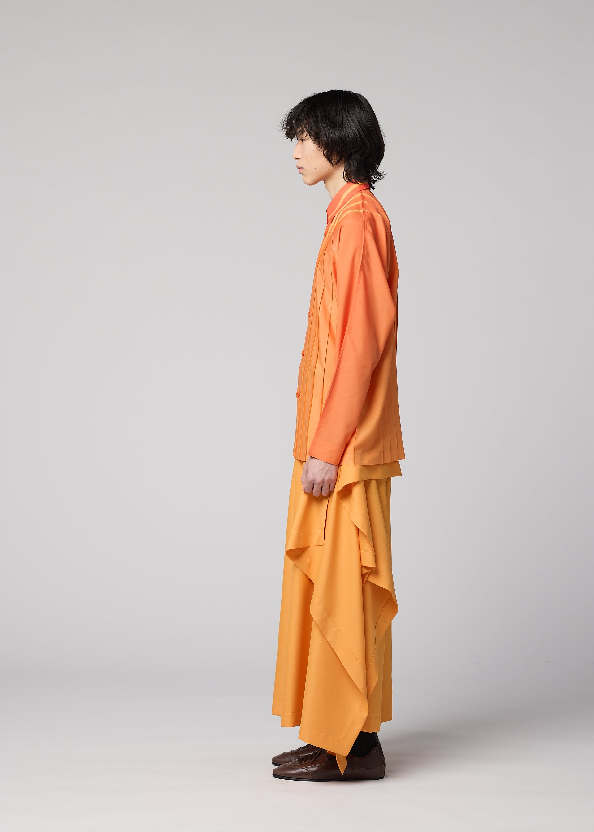 Zoomed view Wearing the IM MEN DAWN RAY SHIRT in orange, a person with medium-length dark hair stands in profile against a plain background, pairing it with a long, flowing yellow skirt featuring a subtle gradation pattern and brown shoes.
