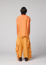 A person stands facing away against a plain gray background, wearing the IM MEN DAWN RAY SHIRT—an orange pleated top with vertical stripes—paired with wide yellow pants featuring draped fabric panels and a subtle gradation pattern.