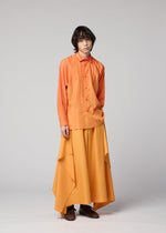 A person with medium-length dark hair is wearing the DAWN RAY SHIRT in orange by IM MEN, paired with a long, flowing yellow skirt featuring draped sides and a subtle gradation pattern, standing against a plain light gray background.