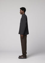 A man in profile stands against a plain light gray background, wearing the IM MEN DOUBLE WEAVE CREPE jacket, brown creased-texture pants, and dark shoes.