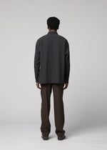 A person with short curly hair stands facing away, wearing an IM MEN DOUBLE WEAVE CREPE dark long-sleeved shirt and brown pants in a plain studio with a light gray background.