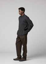 A man wears the IM MEN DOUBLE WEAVE CREPE dark long-sleeved shirt, brown pants, and dark shoes in a studio with a neutral background, looking to the left with his hands in his pockets.