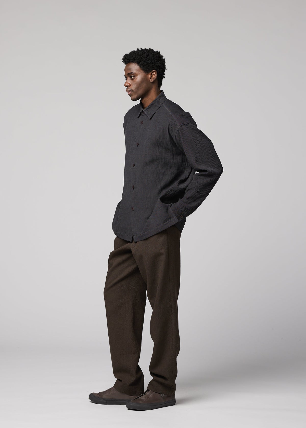 Zoomed view A man wears the IM MEN DOUBLE WEAVE CREPE dark long-sleeved shirt, brown pants, and dark shoes in a studio with a neutral background, looking to the left with his hands in his pockets.