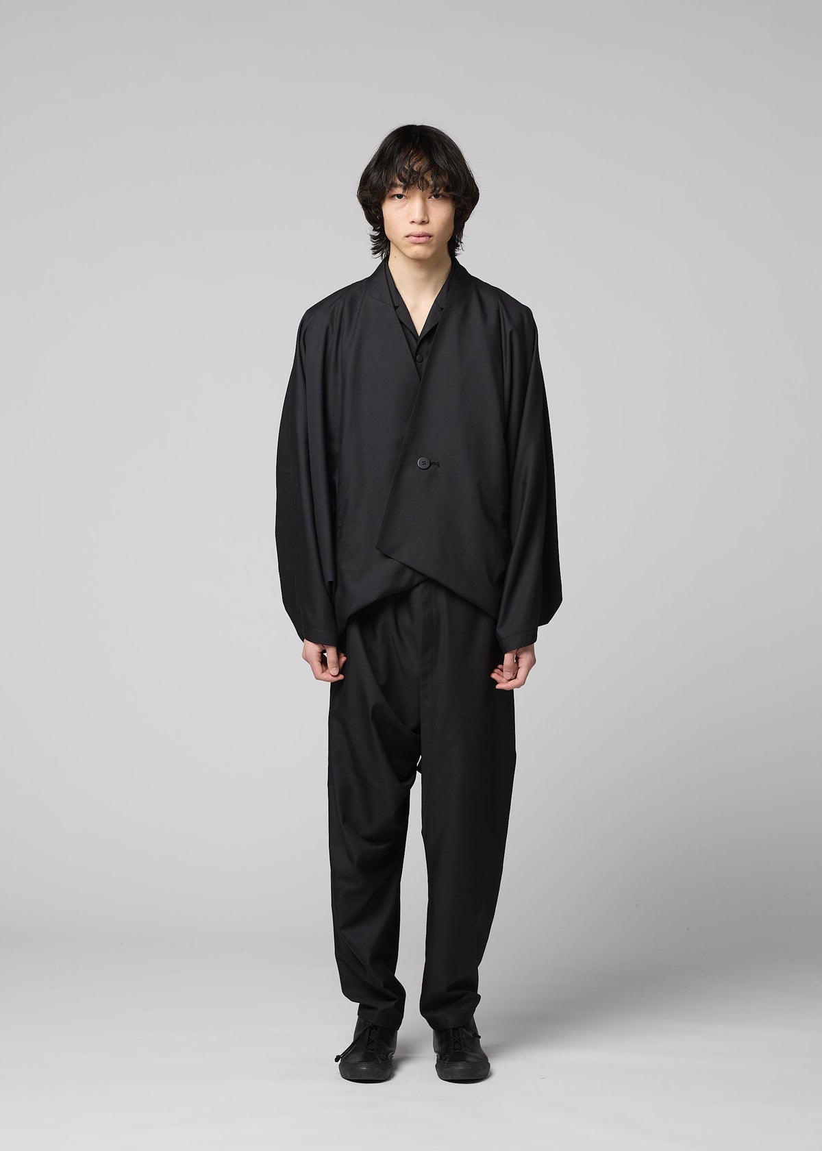 A person with medium-length dark hair stands facing forward against a plain light background, wearing the IM MEN SWING BLACK collarless jacket, paired with black pants and shoes. view 4 of 7