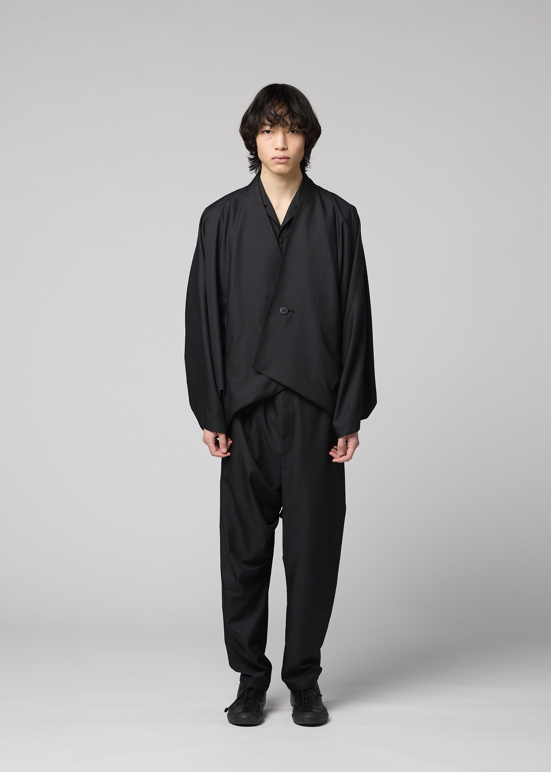 Zoomed view A person with medium-length dark hair stands facing forward against a plain light background, wearing the IM MEN SWING BLACK collarless jacket, paired with black pants and shoes.