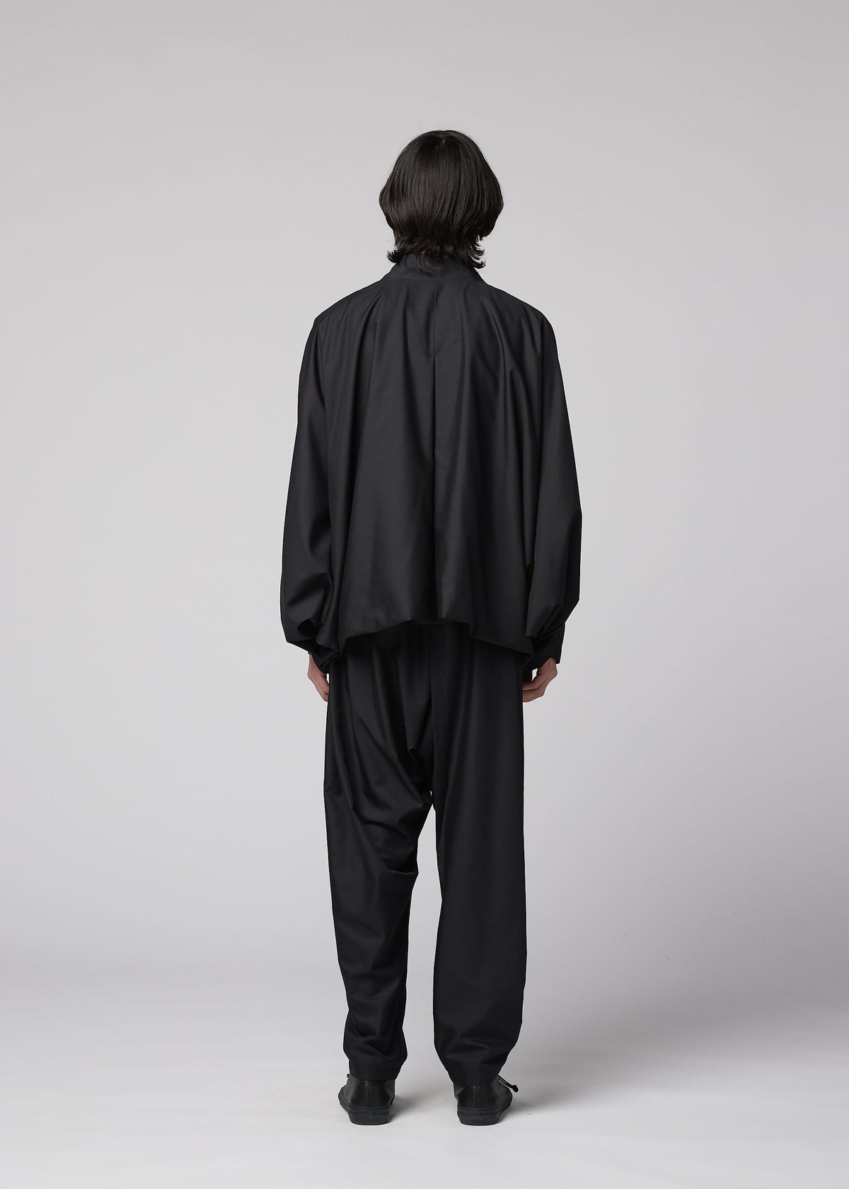 A person with medium-length dark hair faces away, wearing the IM MEN SWING BLACK wrinkle-resistant collarless jacket and matching pants in a minimal studio with a plain light gray background. view 6 of 7