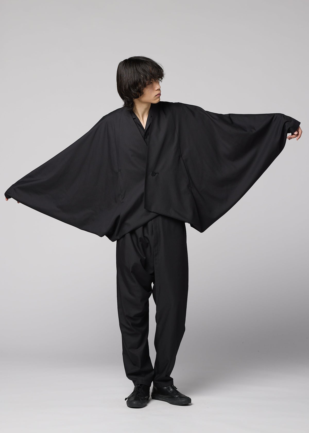 A person in loose, all-black clothing poses with arms extended, highlighting the wide, flowing SWING BLACK jacket by IM MEN, reminiscent of bat wings against a plain light background. view 7 of 7