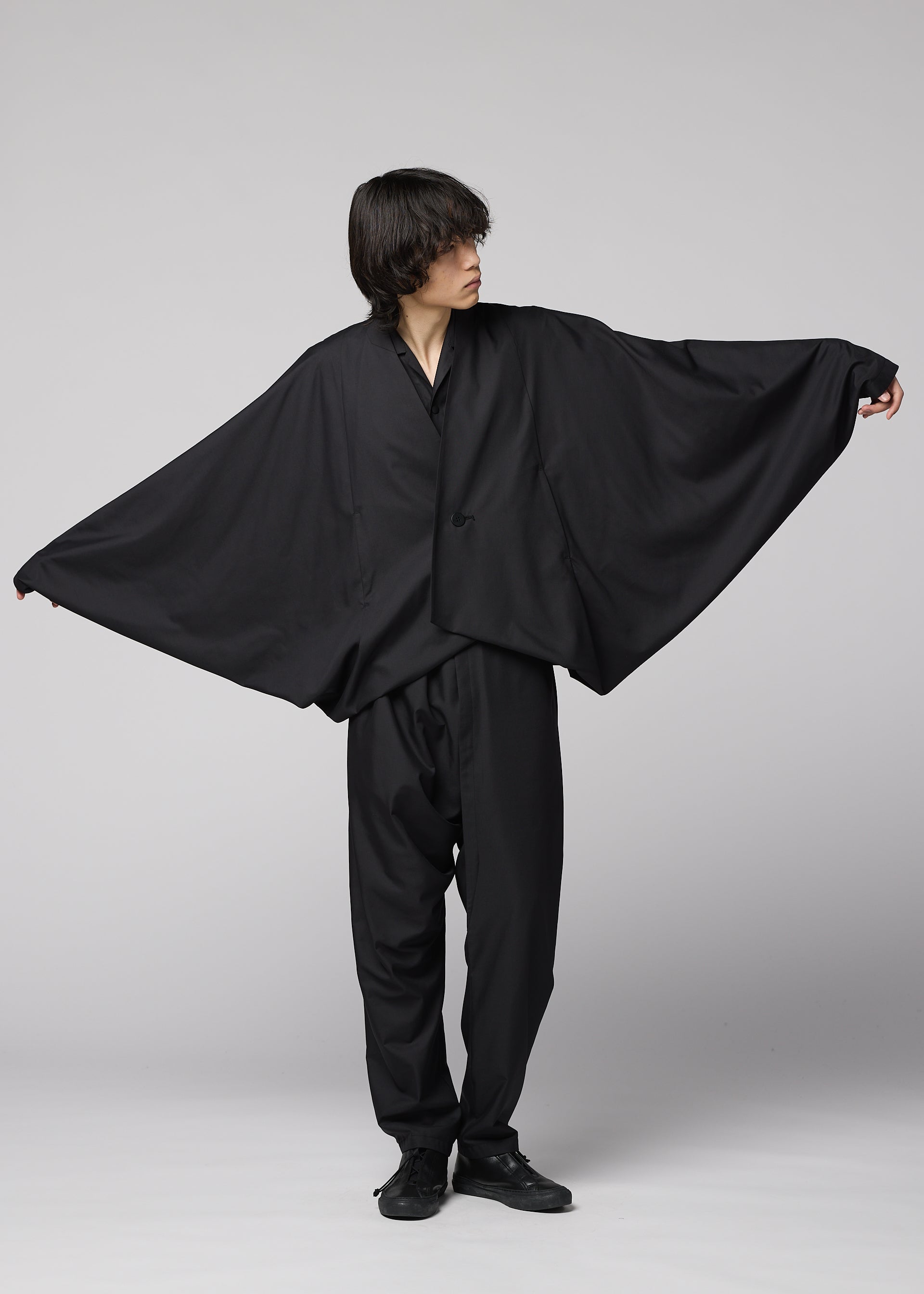 Zoomed view A person in loose, all-black clothing poses with arms extended, highlighting the wide, flowing SWING BLACK jacket by IM MEN, reminiscent of bat wings against a plain light background.