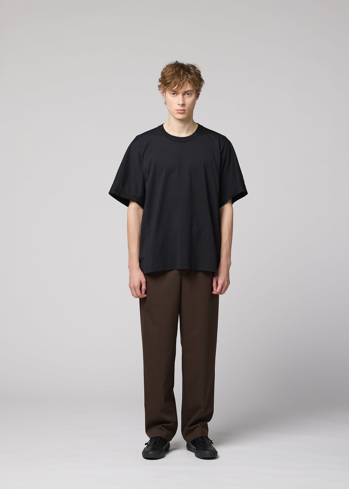 A person with short, tousled hair stands facing forward, wearing IM MEN’s TURN loose black jersey T-shirt with 3D construction, brown pants, and black shoes against a light gray background. view 5 of 8