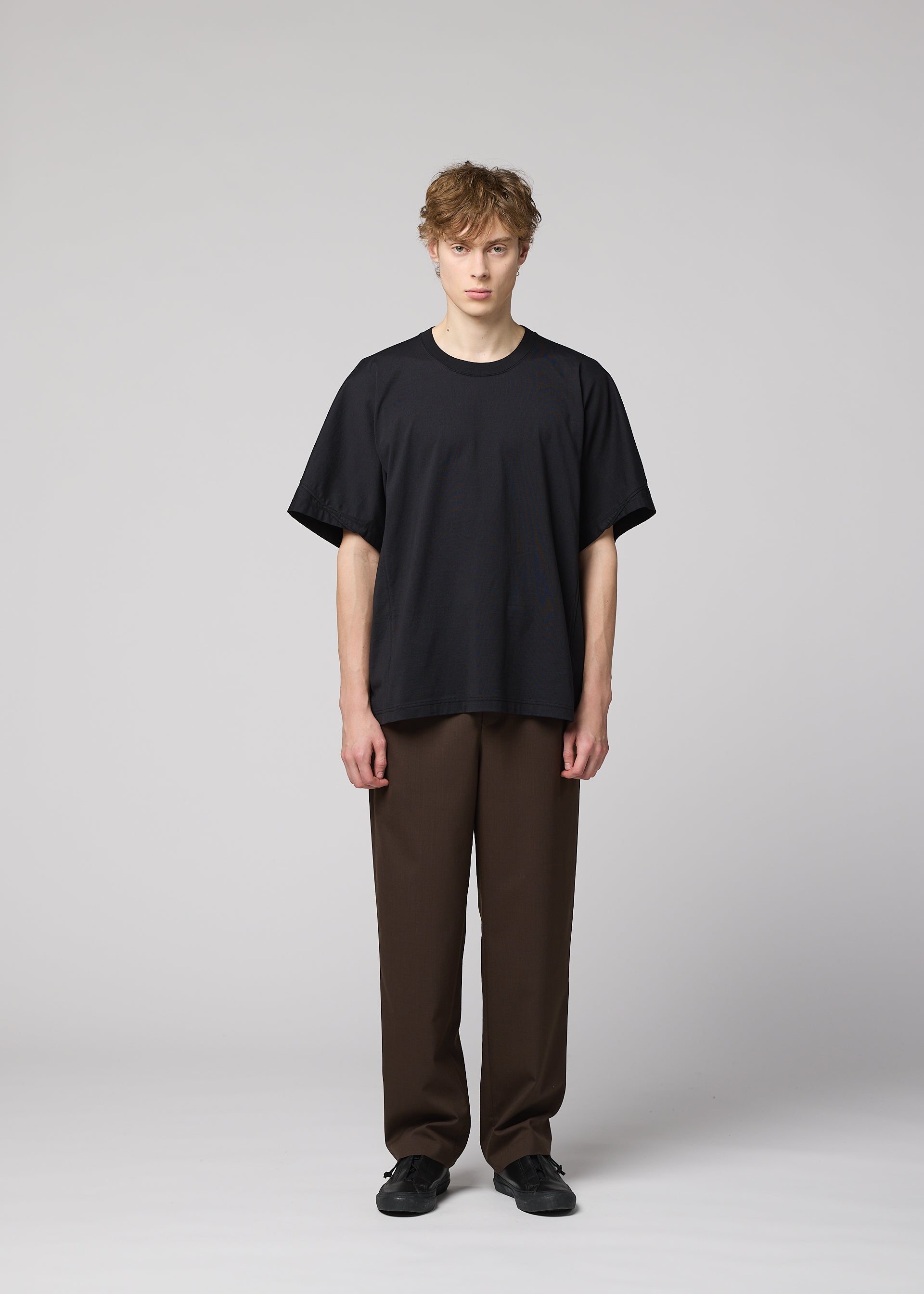 Zoomed view A person with short, tousled hair stands facing forward, wearing IM MEN’s TURN loose black jersey T-shirt with 3D construction, brown pants, and black shoes against a light gray background.