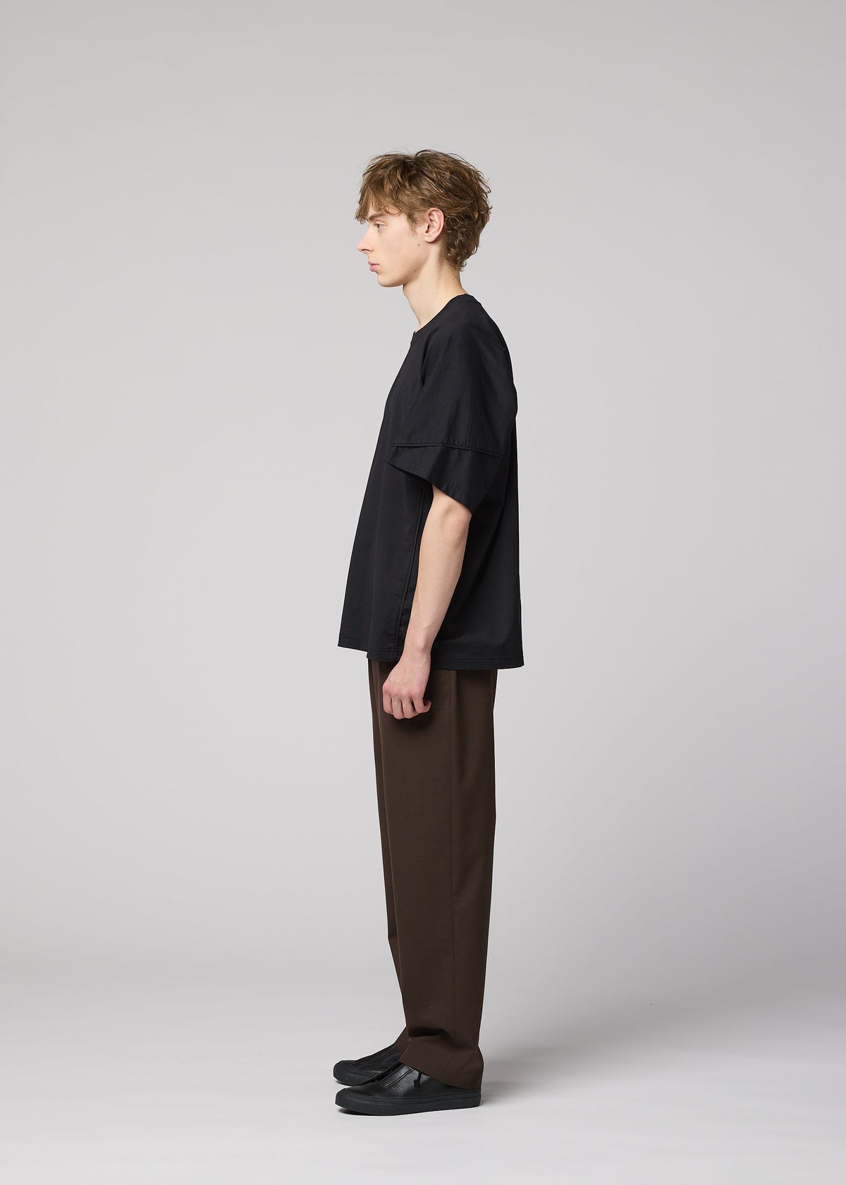 A young person with short light brown hair stands in profile against a gray background, wearing the IM MEN TURN loose black jersey T-shirt, paired with brown pants and black shoes. view 6 of 8