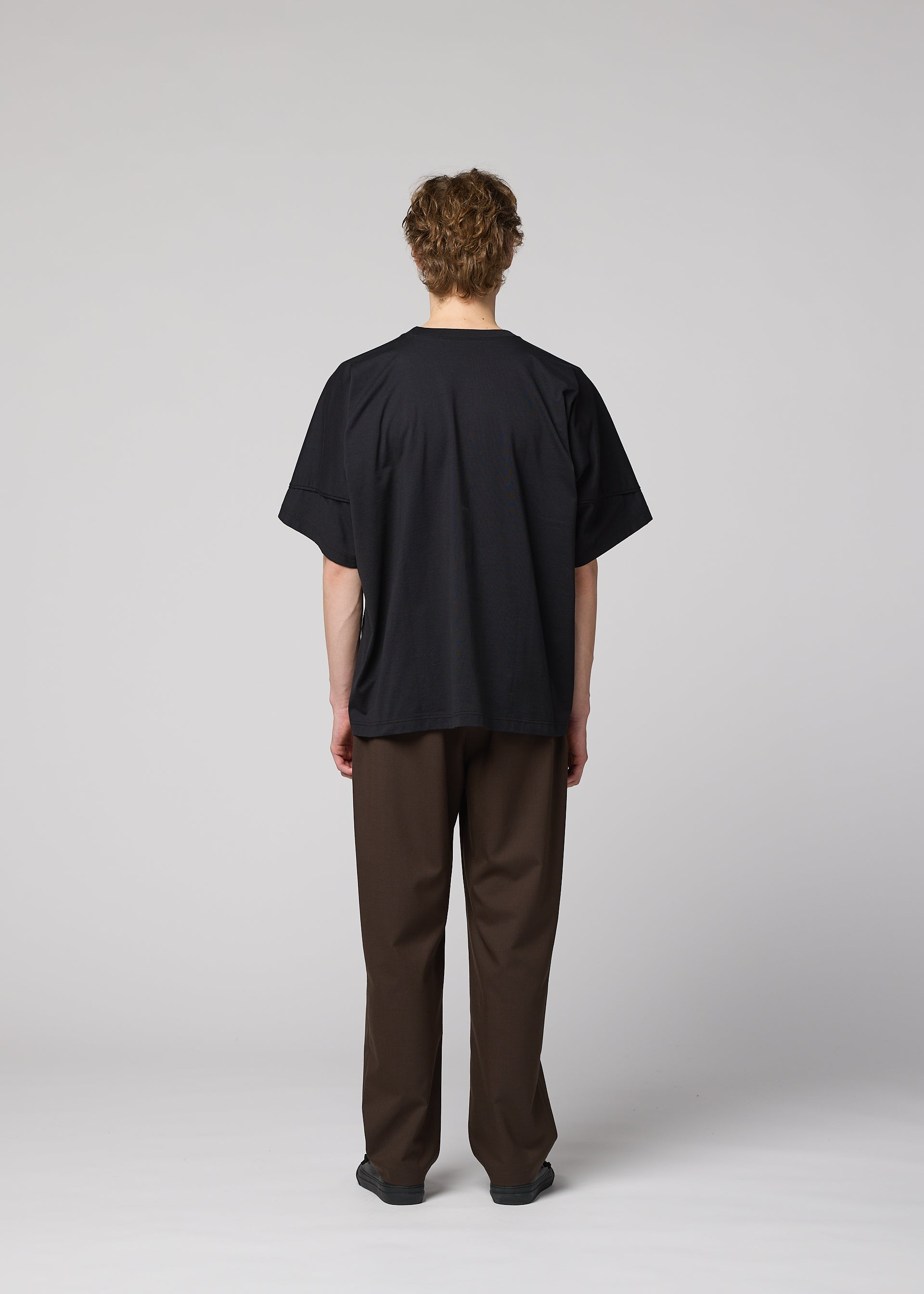 Zoomed view A person with short curly hair faces away from the camera, wearing the IM MEN TURN loose black jersey T-shirt, brown pants, and black shoes against a plain light gray background.