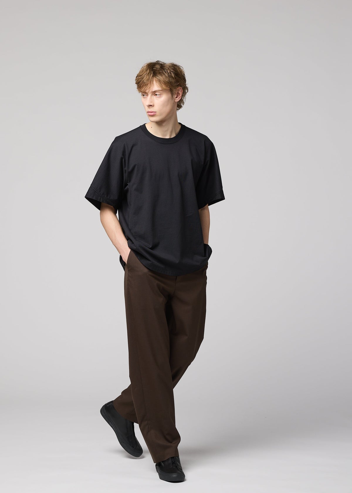 A young man with light brown hair wears IM MEN’s TURN: a loose black jersey T-shirt, wide brown trousers, and black shoes. Hands in pockets, he walks against a plain light gray background, embodying modern simplicity. view 8 of 8